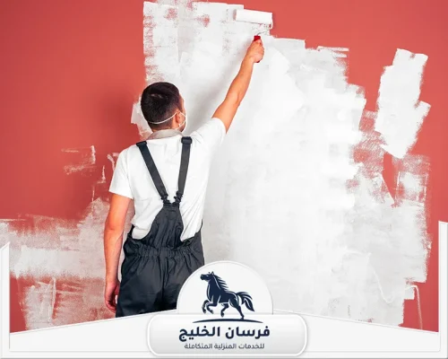 خدمات صباغ في الفجيرة - فرسان الخليج فني دهانات يوزع الطلاء الأبيض على جدار أحمر باستخدام أسطوانة الطلاء.