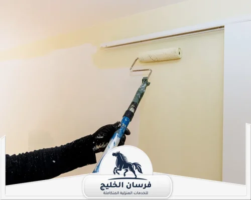 شركة صبغ في الشارقة - فرسان الخليج رول دهان طويل يوزع طلاءً بيج فاتحًا على جدار داخلي بجوار إطار نافذة.
