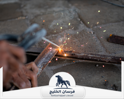حداد يلحم قطعتين من الحديد باستخدام جهاز لحام كهربائي داخل الورشة.