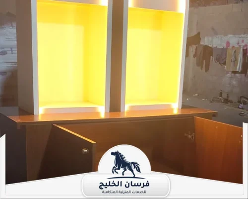 تفصيل اثاث في عجمان - فرسان الخليج وحدة أثاث خشبية تضم خزائن سفلية ورفوف عرض علوية بإضاءة صفراء.