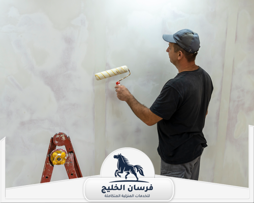 معلم صباغ في أم القيوين 0547761153 فرسان الخليج عامل صباغة يغطي الحائط البرتقالي بطبقة طلاء رمادية.
