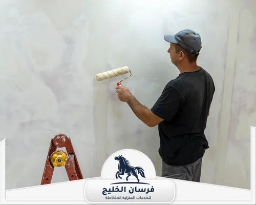 معلم صباغ في أم القيوين 0547761153 فرسان الخليج عامل صباغة يغطي الحائط البرتقالي بطبقة طلاء رمادية.