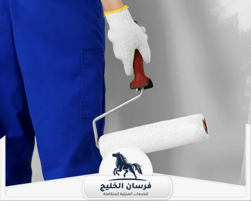 صباغ في راس الخيمة - فرسان الخليج عامل دهانات يرتدي زيًا أزرق ويحمل أسطوانة طلاء بجوار جدار داخلي.