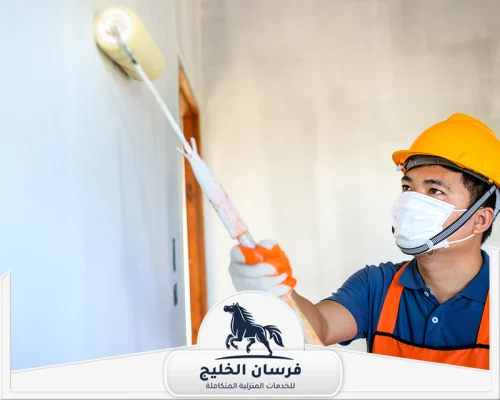 شركة ديكور داخلي في عجمان - فرسان الخليج فني دهانات يطلي الجدار بالرول مع ارتداء خوذة وكمامة وقفازات واقية.