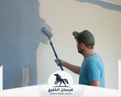 معلم دهان في رأس الخيمة - فرسان الخليج عامل يرتدي قفازات أثناء تطبيق الدهان الرمادي على جدران المنزل.