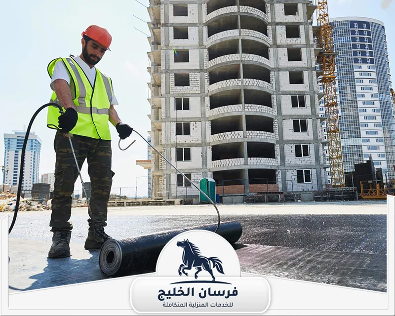 اسعار شركة عزل اسطح ابوظبي - فرسان الخليج عامل يطبق العزل البيتوميني على سطح مبنى باستخدام معدات متخصصة.