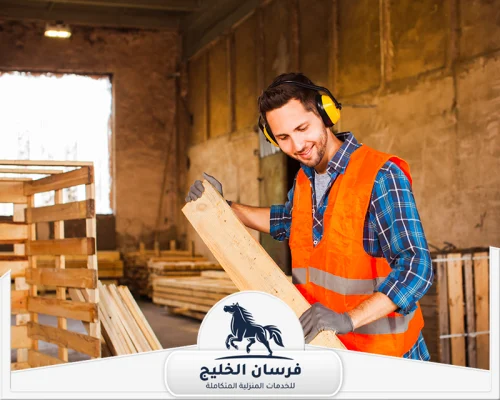 نجار خشب طاولات في عجمان 0547761153 فرسان الخليج نجار يحمل لوح خشب داخل الورشة استعدادًا لأعمال القص والتجهيز.