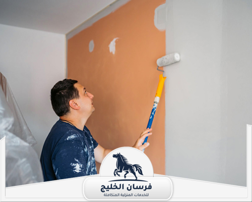صباغ جدران في أم القيوين 0547761153 فرسان الخليج عامل صباغة يغطي الحائط البرتقالي بطبقة طلاء رمادية.