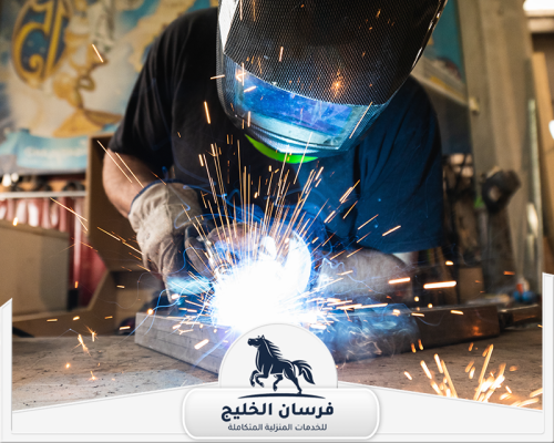 حداد شبابيك في عجمان 0547761153 فرسان الخليج فني لحام يرتدي قناع الحماية أثناء تثبيت قطع حديدية وسط تطاير الشرر.