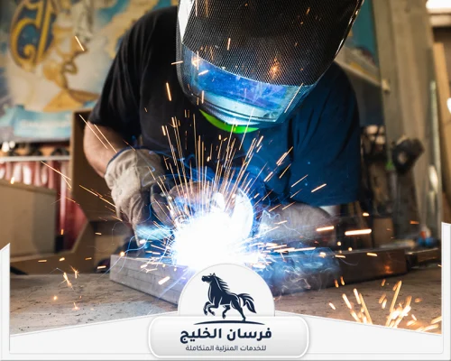 حداد شبابيك في عجمان 0547761153 فرسان الخليج فني لحام يرتدي قناع الحماية أثناء تثبيت قطع حديدية وسط تطاير الشرر.
